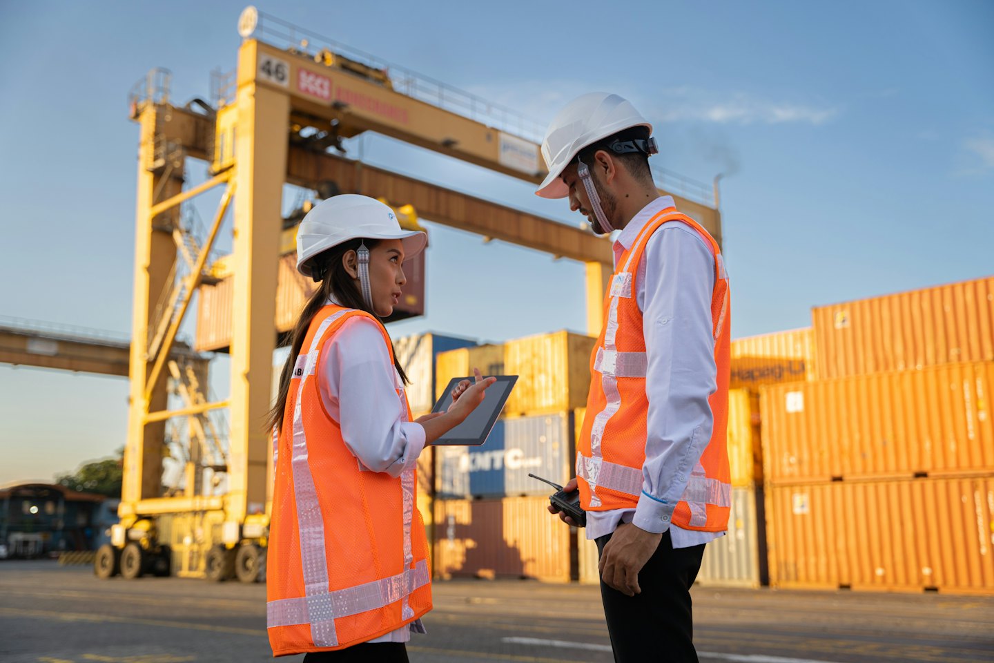 Två logistikarbetare diskuterar hamnverksamhet med containrar och kranar.